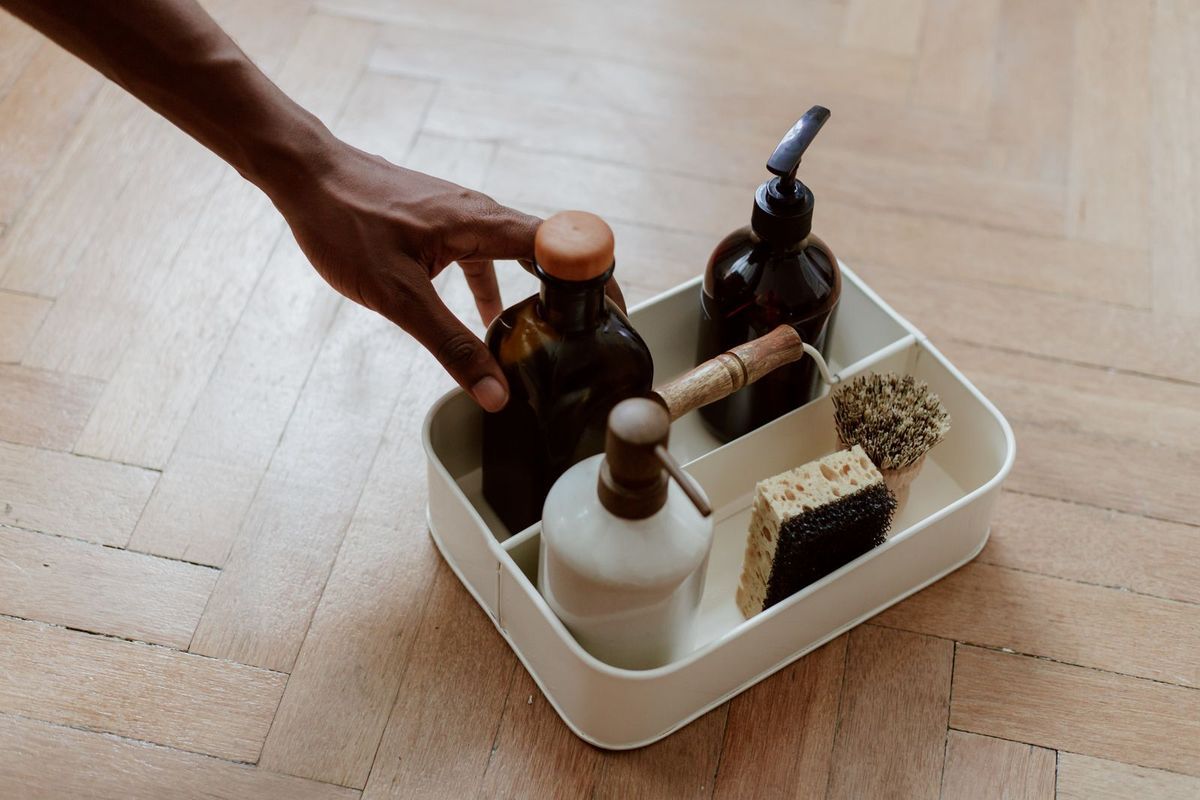 Hand reaching into a caddy of cleaning bottles, a sponge and a brush on a wood floor