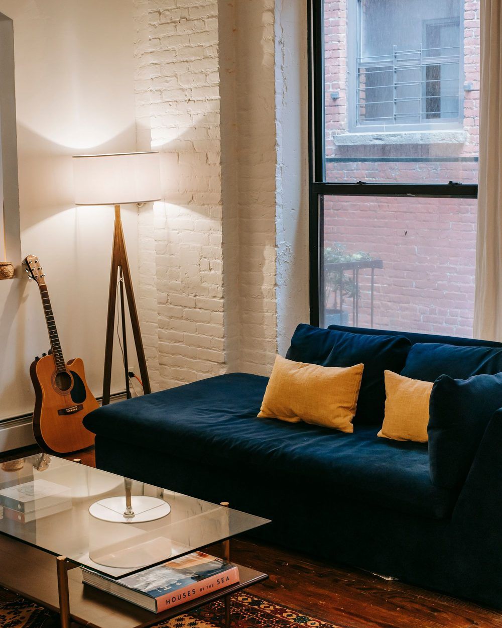 Navy sofa with yellow pillows, a floor lamp and a guitar in a sunny apartment living room with a brick wall