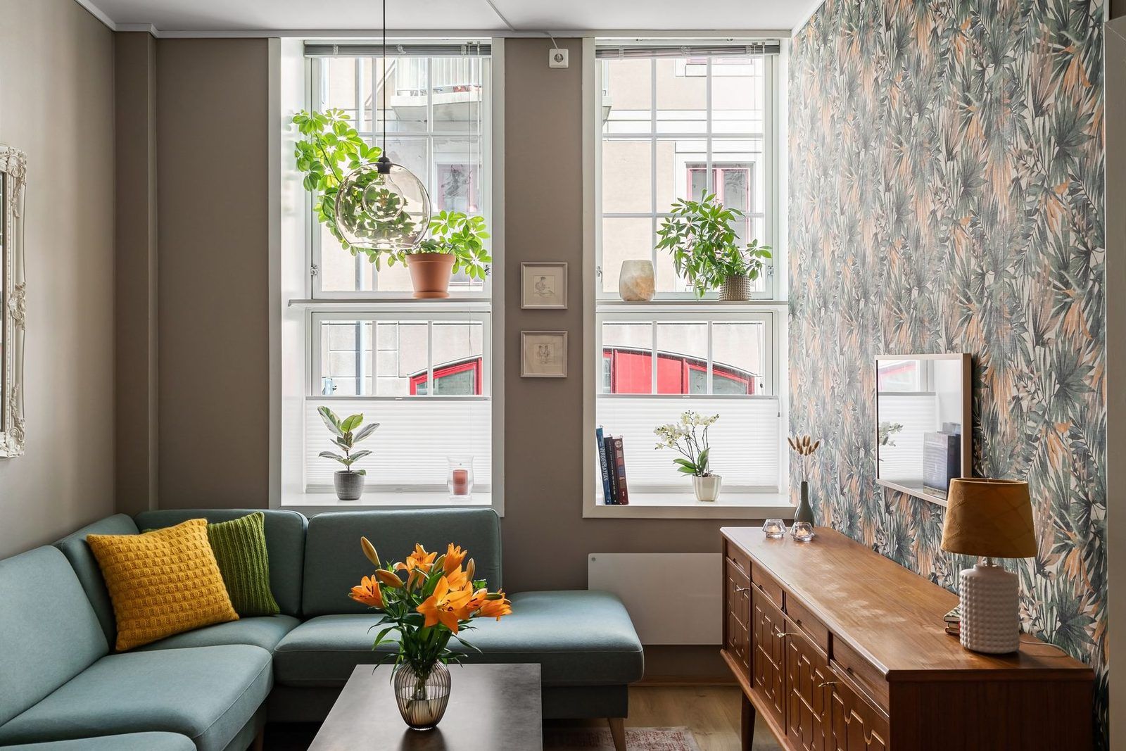 Bright living room with a teal sectional sofa, plants on the windowsills and a leaf-patterned accent wall
