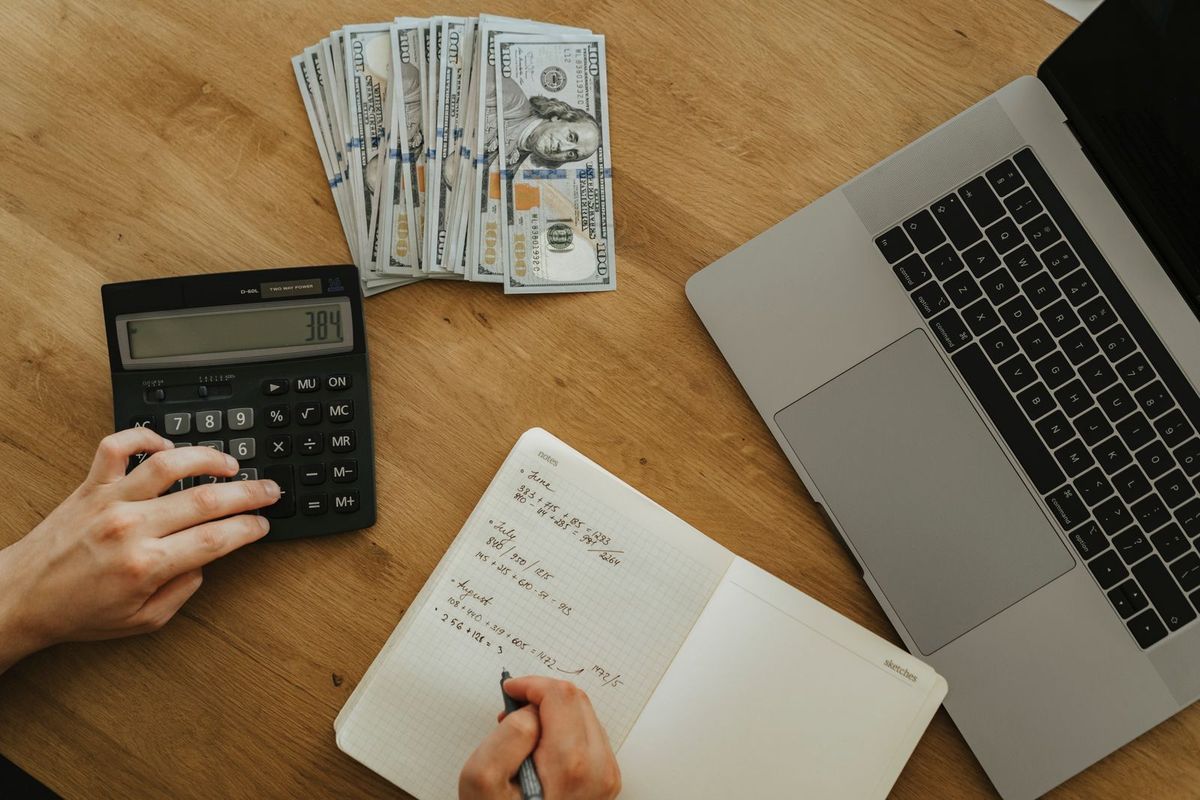 Hands using a calculator and writing sums in a notebook next to a laptop and dollar bills