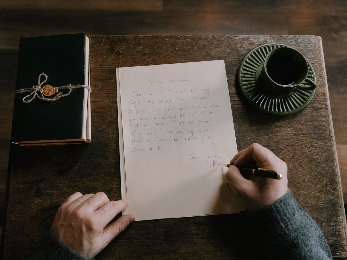 Hands writing a letter at a wooden desk next to a cup of coffee